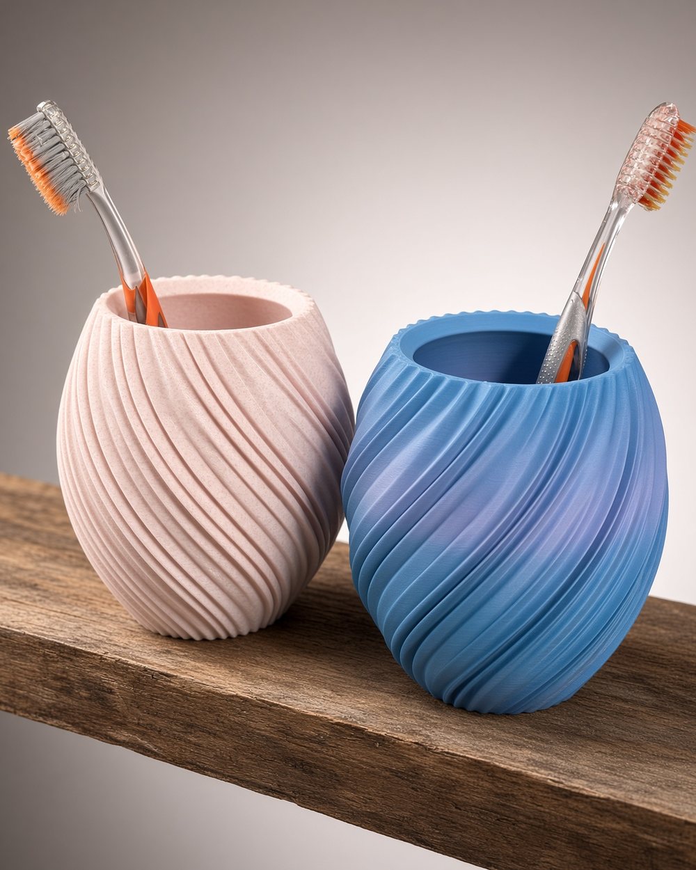 A pink and a blue spiral toothbrush cup side by side on a bathroom shelf, a toothbrush in each