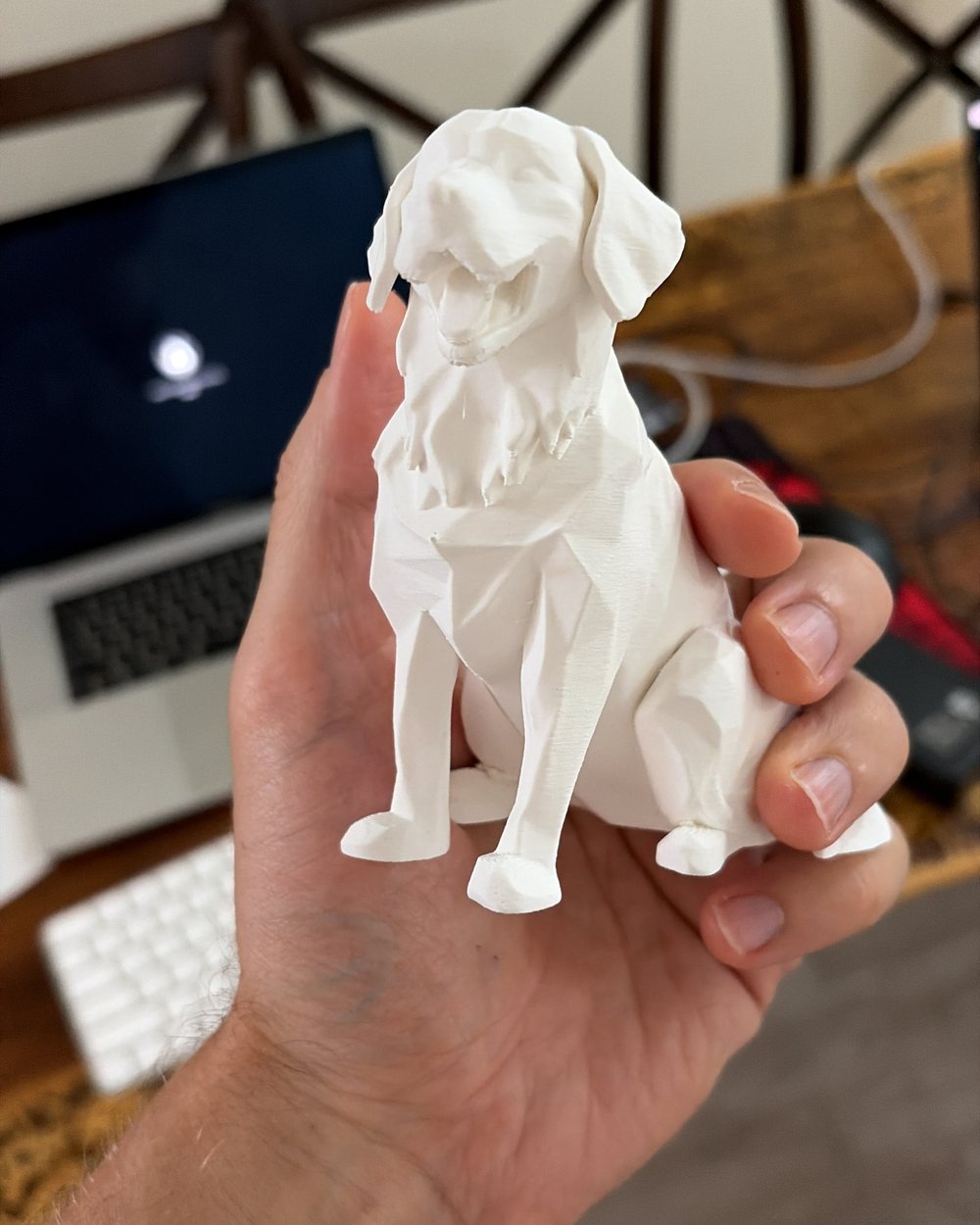 A white low-poly dog sitting, held in a hand in front of a desk