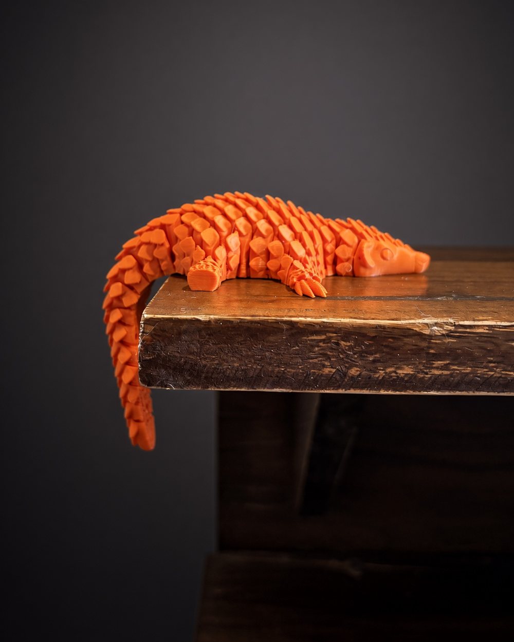 An orange articulated pangolin on the edge of a wooden table, its tail hanging over the side