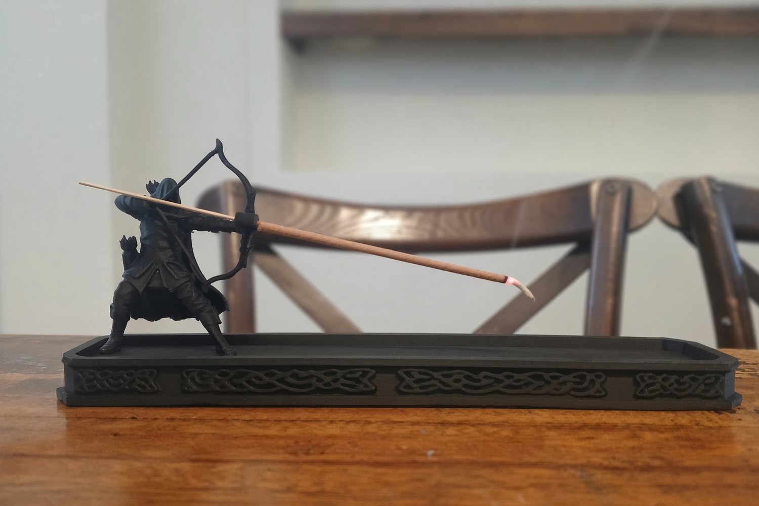 A black printed archer kneeling at one end of a long incense tray, drawing his bow; the incense stick is the arrow and smoke rises from its lit tip