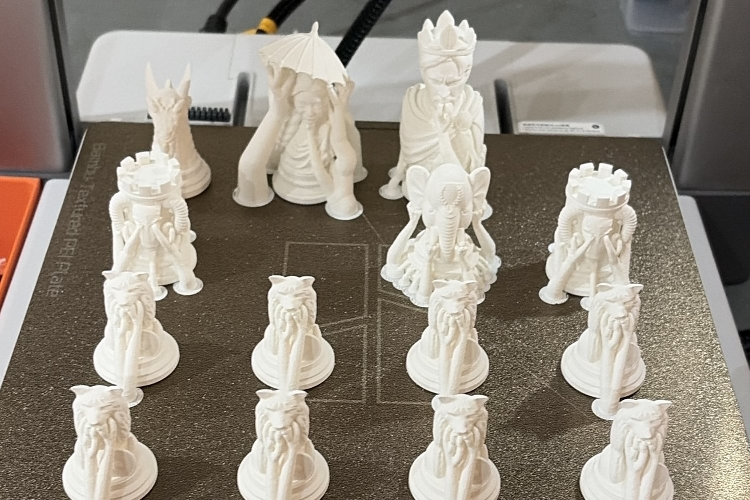 The printed white chess pieces standing on the printer's build plate, the queen under her parasol among them